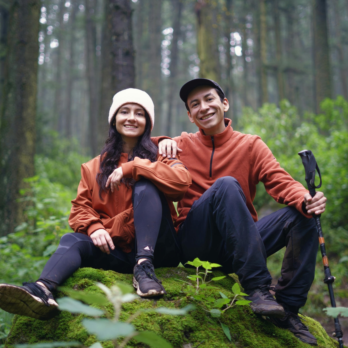 Happy couple in orange jackets resting on mossy log during forest hiking adventure