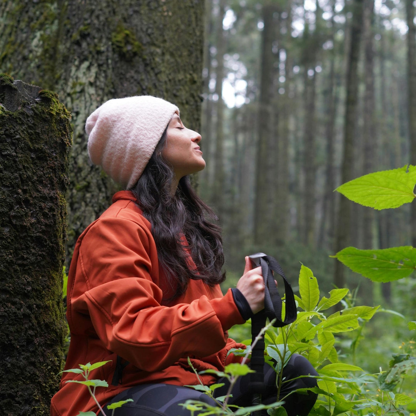 Woman in a forest, sitting against a tree, holding a camera.