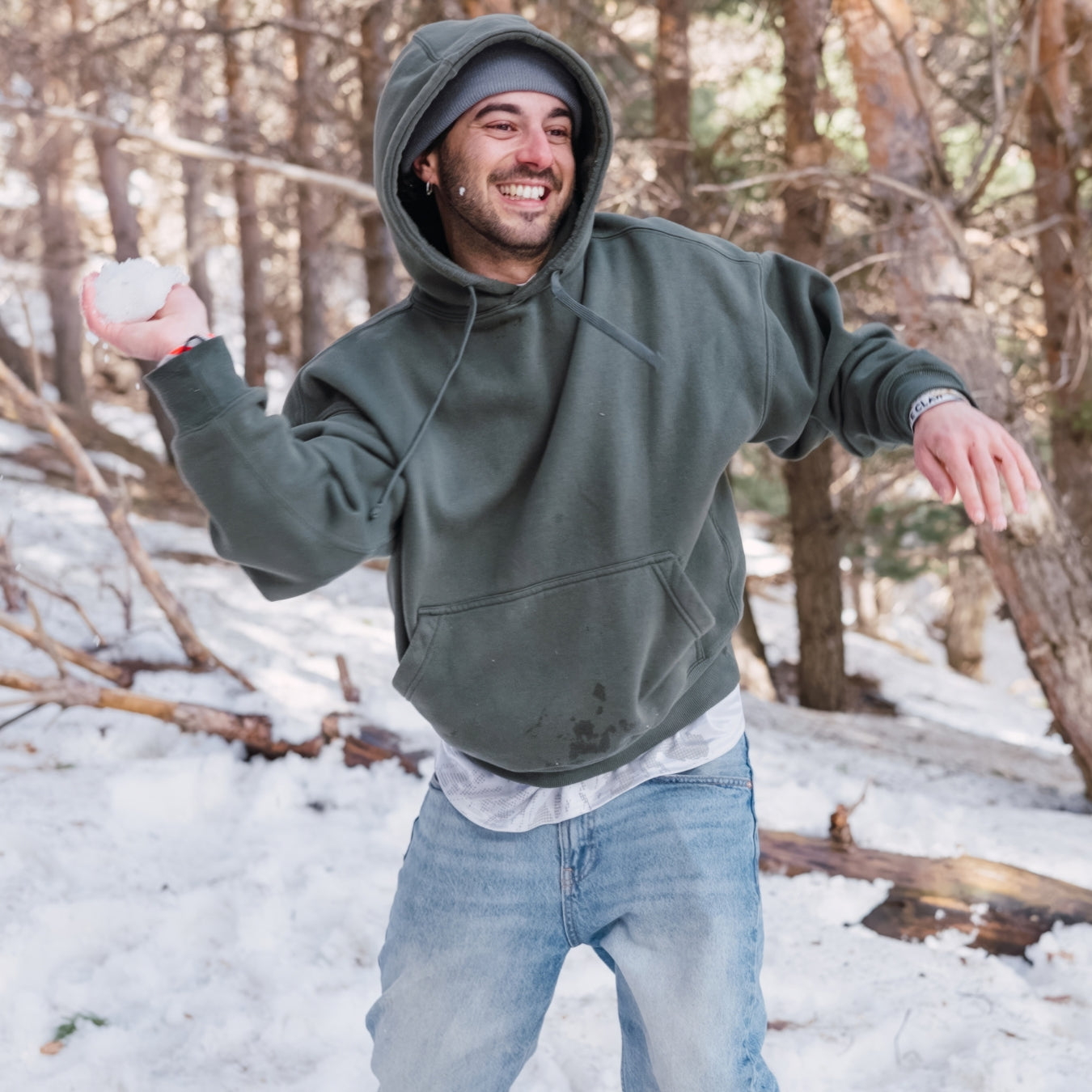 Man in a gray hoodie and jeans standing in a snowy forest, holding a snowball.